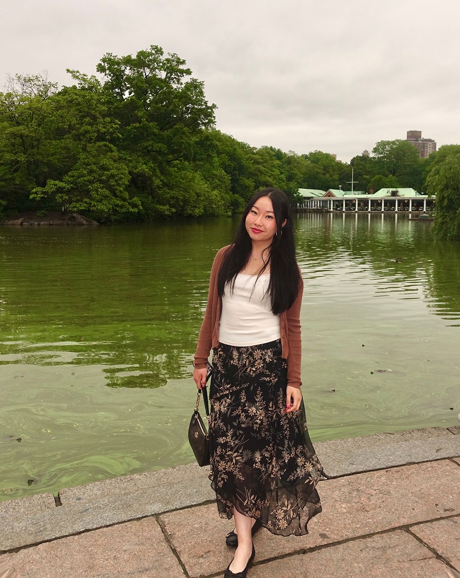 Portrait of Tina Zou standing beside a lake in a park, wearing a floral skirt and cardigan.
